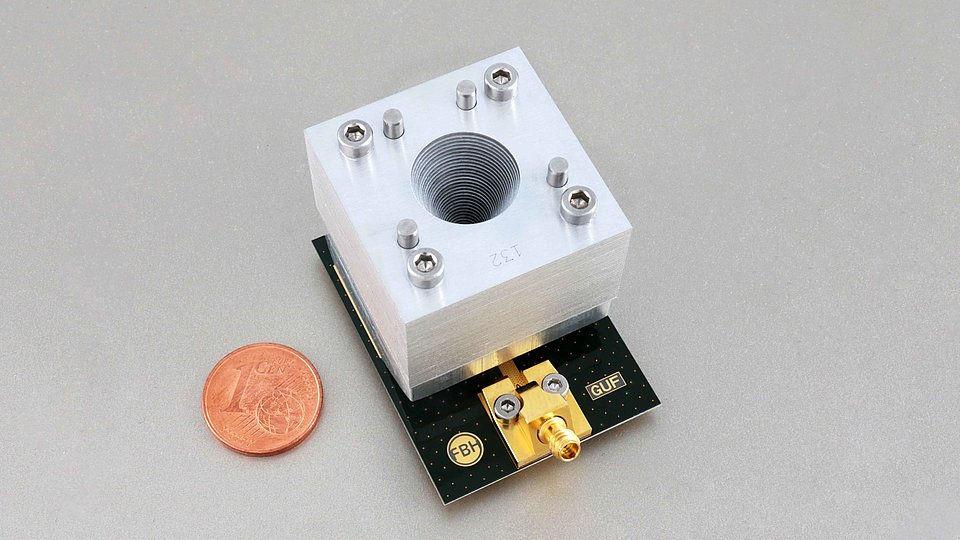 A close-up of a metallic waveguide component with a cylindrical hole, mounted on a circuit board labeled "FBH" and "GUF." A one-cent euro coin is placed nearby for size comparison.