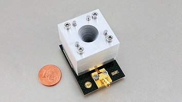 A close-up of a metallic waveguide component with a cylindrical hole, mounted on a circuit board labeled "FBH" and "GUF." A one-cent euro coin is placed nearby for size comparison.