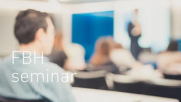 Publikum sitzt und blickt auf einen unscharfen Vortragenden, der vorne in einem Raum spricht, mit eingeblendetem Text „FBH Seminar“.