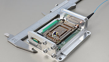Lasermodul neben einem Messschieber, der verdeutlicht, dass das Modul nicht größer als 10 cm ist. Im Modul sieht man verschiedene Linsen und einen Kristall. Von außen ist das Modul mit einer Faser gekoppelt, die die Pumpstrahlung in das Modul leitet.
