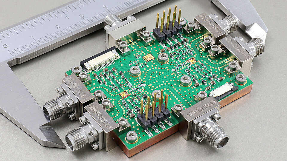 Eine grüne Leiterplatte mit verschiedenen Komponenten, wie Widerständen und Verbindungsstellen, liegt auf einem Tisch. Links und rechts sind Metallanschlüsse sichtbar. Eine digitale Schieblehre misst die Größe der Leiterplatte.