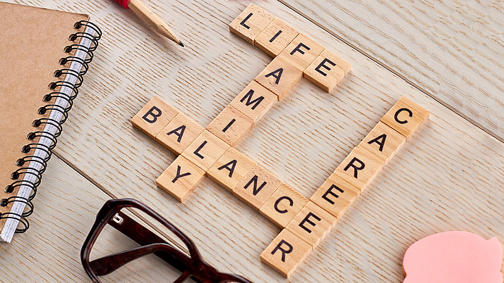 Wooden letter tiles arranged to spell “LIFE,” “BALANCE,” “FAMILY,” and “CAREER” in a crossword style. Surrounding elements include a notebook and a pair of glasses.