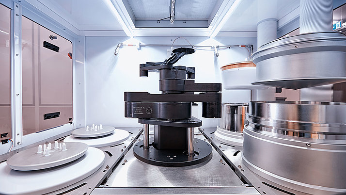 Moderne Maschinenanordnung in einem Reinraum. Im Zentrum befindet sich ein schwarzer, drehbarer Roboterarm, der präzise mit verschiedenen runden Metall- und Kunststoffplatten arbeitet. 