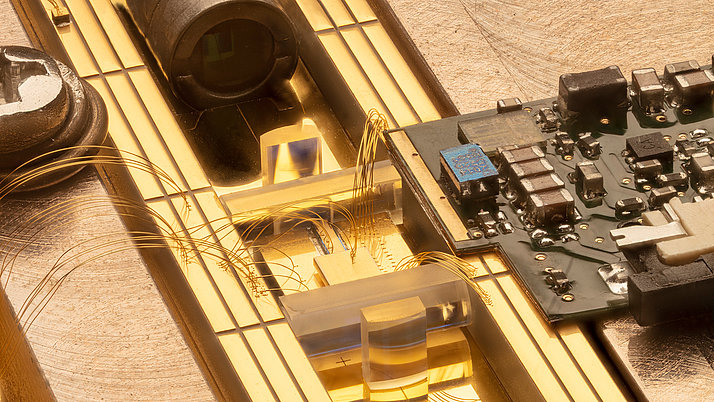 Detailaufnahme einer elektronischen Schaltung, die aus einer Platine und klaren, prismatischen Bauteilen besteht. Feine Drähte verbinden die Komponenten, während ein goldener Untergrund die Struktur unterstützt.
