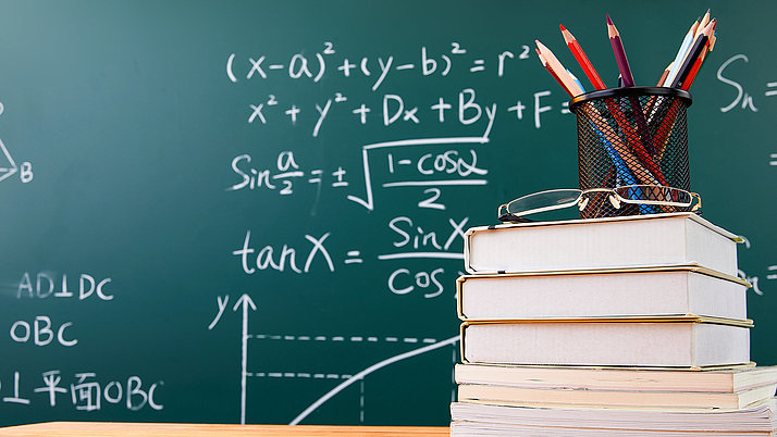 Symbolic image: in the background a blackboard written with formulas. In front of it a stack of books with pens on top of it 