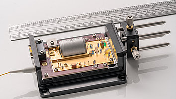 Open quantum light laser module with fiber connection, showing a gold-colored circuit board inside with various optical components, a large silver element, transparent lenses, and other microelectronic components. A silver-colored ruler is placed in the background of the image for size comparison.