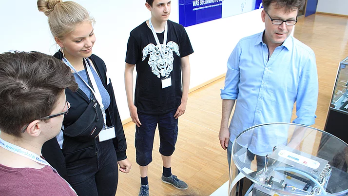 Eine Gruppe von drei Jugendlichen und einem Erwachsenen steht um einen Glaskasten herum und schaut auf ein technisches Gerät. Der Erwachsene erklärt etwas zu dem Modell, während die Jugendlichen aufmerksam zuhören.