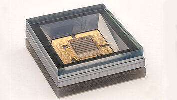 A transparent microchip in a rectangular housing mounted on a golden circuit board. The chip shows fine lines and structures. 