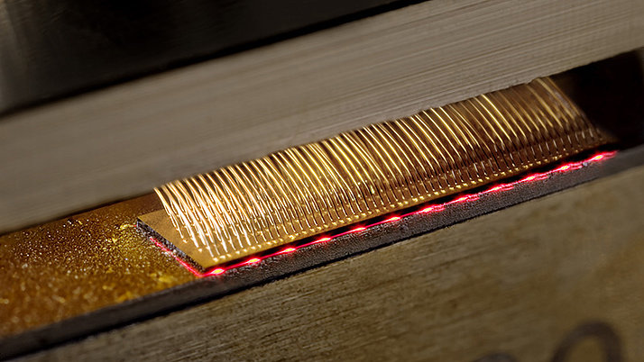 Close-up of a gold-colored wire visible in a device surrounded by a red line of light. The wire is positioned in a gap between two pieces of wood lying on top of each other.