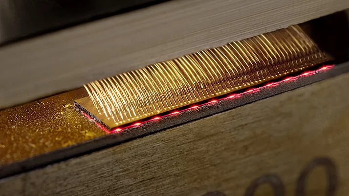 Close-up of a gold-colored wire visible in a device surrounded by a red line of light. The wire is positioned in a gap between two pieces of wood lying on top of each other.