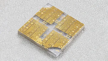 Photograph of a small square semiconductor wafer with multiple gold-colored transistor device patterns arranged in four sections on a transparent substrate.
