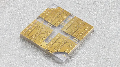 Photograph of a small square semiconductor wafer with multiple gold-colored transistor device patterns arranged in four sections on a transparent substrate.