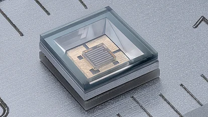 Close-up of a square LED chip with parallel metallic strips, mounted on a gold-colored base inside a recessed metallic package with a transparent cover.