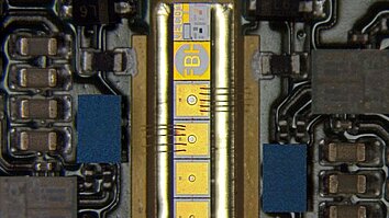 Microscope image of an electronic device package with a narrow central chip connected by multiple wire bonds to surrounding circuitry. Gold contact pads and printed markings are visible on the chip, with surface-mounted components arranged along both sides.