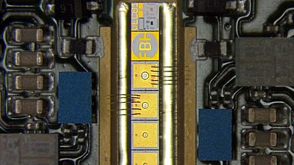 Microscope image of an electronic device package with a narrow central chip connected by multiple wire bonds to surrounding circuitry. Gold contact pads and printed markings are visible on the chip, with surface-mounted components arranged along both sides.
