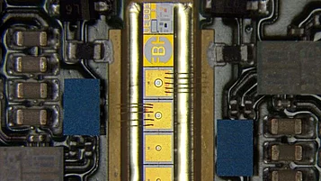 Microscope image of an electronic device package with a narrow central chip connected by multiple wire bonds to surrounding circuitry. Gold contact pads and printed markings are visible on the chip, with surface-mounted components arranged along both sides.