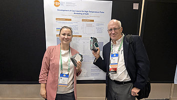 Ina Ostermay (left) from the FBH stands next to Marty Brophy (chair of the poster session) in front of a research poster during the CS Mantech Conference. They are holding samples in their hands and smiling. The poster describes the development of cap layers for high-temperature annealing of GaN. The FBH logo is visible in the background.