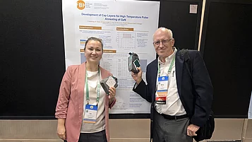 Ina Ostermay (links) aus dem FBH steht neben Marty Brophy (Chair der Postersession) vor einem Forschungsposter. während der CS Mantech Konferenz Sie halten Proben in der Hand und lächeln. Das Poster beschreibt die Entwicklung von Cap-Sschichten für das Hochtemperatur-Annealing von GaN. Das Logo des FBH ist im Hintergrund sichtbar.