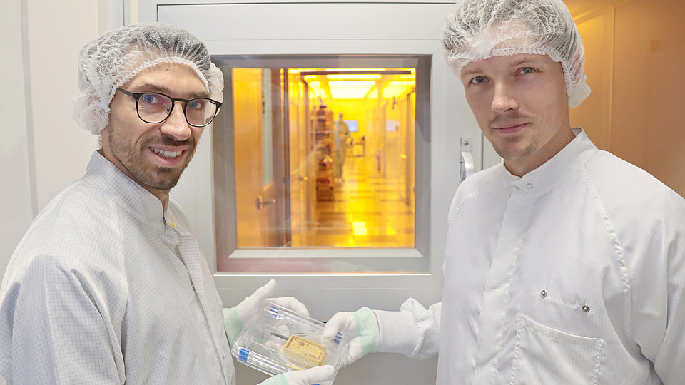 Two employees hold a micromodule in their hands in front of the entrance to the cleanroom and look into the camera.