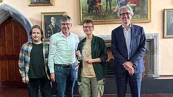 Vier Personen bei einer Preisverleihung; die ausgezeichnete Person in der Mitte hält eine Glastrophäe.