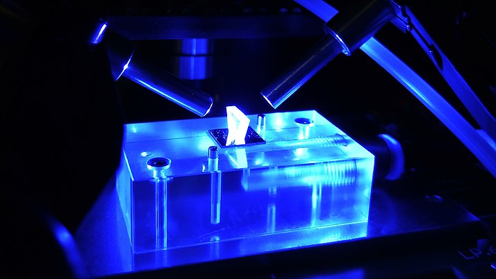 An enlarged view of a transparent object, illuminated in blue, between two microscopes. It shows a crystal-like structure on a flat surface.