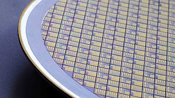 Halbleiterwafer mit regelmäßig angeordneten, goldenen und violetten Linien und Mustern. Der Wafer hat einen runden Durchmesser und ist auf einem dunklen Hintergrund platziert.