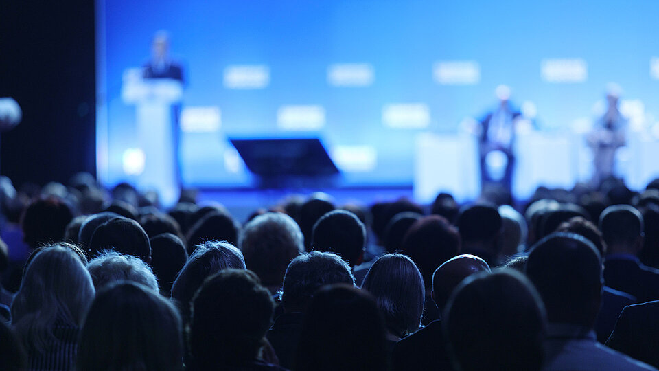 Große Menschenmenge sitzt und hört einer Rede auf einer Bühne mit blauem Hintergrund bei einer Konferenz oder Veranstaltung zu.