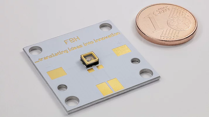 An electronic circuit board with gold and silver elements is shown in the foreground, with a small sensor in the middle. To the right of it is a 1-cent coin. On the circuit board is the lettering “FBH - translating ideas into innovation”.