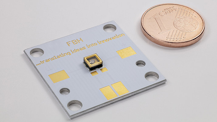 An electronic circuit board with gold and silver elements is shown in the foreground, with a small sensor in the middle. To the right of it is a 1-cent coin. On the circuit board is the lettering “FBH - translating ideas into innovation”.