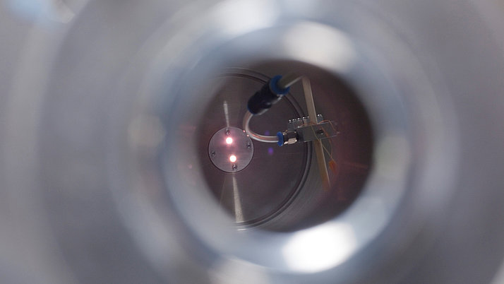 Blick durch ein Rohr auf einen metallischen Zylinder mit einer runden Fläche in der Mitte. Dort sind zwei kleine Lichtquellen sichtbar, die schwach leuchten. 