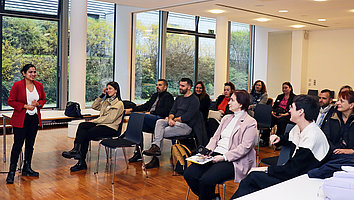 A woman in a red blazer is giving a presentation to a small group of attendees in a modern seminar room with large windows. The audience is seated on chairs and listening attentively.
