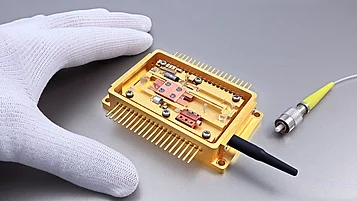 Goldfarbenes offenes Gehäuse mit Glasfaser-Anschluß. Im Inneren des Moduls befindet sich auf einem gefrästen Inlay der Halbleiterlaser, ein Mikroisolator, Umlenkspiegel, ein nicht-linearer Kristall zur Frequenzverdopplung sowie die Faserkopplung.