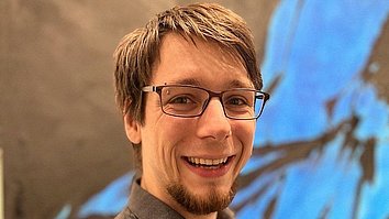 A smiling man with glasses stands in front of an abstract, colorful wall. He is wearing a dark, polka-dotted shirt and has short, light brown hair.