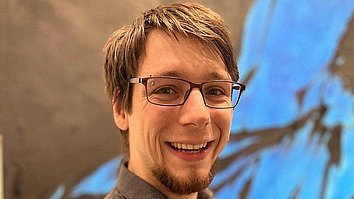 Ein lächelnder Mann mit Brille steht vor einer abstrakten, farbenfrohen Wand. Er trägt ein dunkles, gepunktetes Hemd und hat kurzes, hellbraunes Haar.