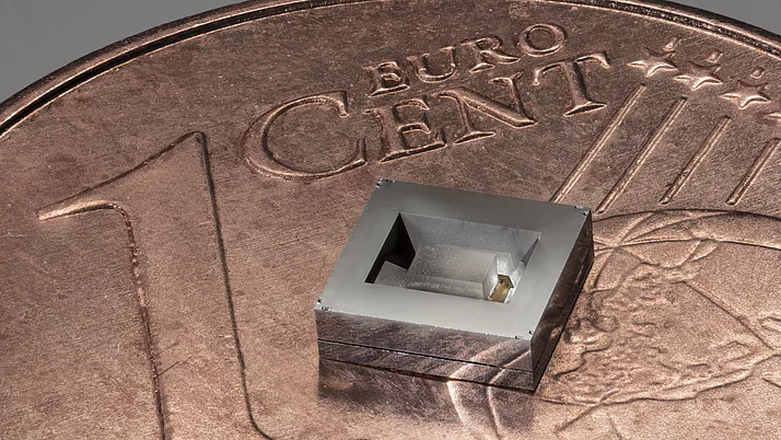 Ein quadratisches, silbernes Mikrochip-Design liegt auf einer 1-Cent-Münze. Der Chip hat eine eingekerbte Form in der Mitte, die seine Struktur sichtbar macht. 