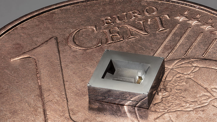 Ein quadratisches, silbernes Mikrochip-Design liegt auf einer 1-Cent-Münze. Der Chip hat eine eingekerbte Form in der Mitte, die seine Struktur sichtbar macht. 