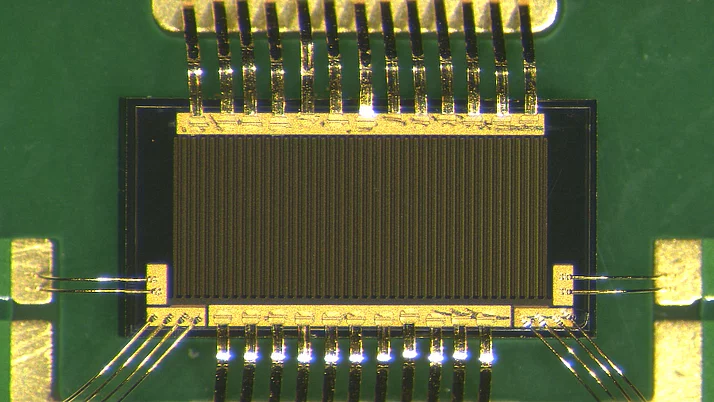 Mikrochip mit goldenen Anschlussdrähten, die an eine grüne Leiterplatte verbunden sind. Der Chip hat eine rechteckige Form und ist mit vertikalen Streifen versehen, die seine Struktur bilden.