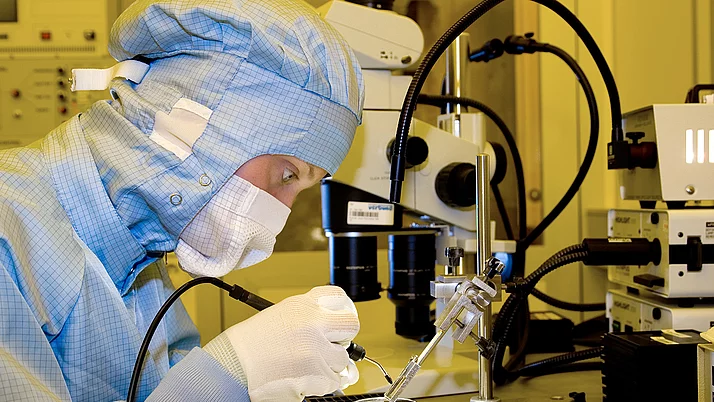 Eine Person in einem sauberen Raum trägt eine Schutzkleidung und arbeitet unter einem Mikroskop. Sie hält ein feines Werkzeug in der Hand, während sie an einem kleinen Objekt in einem Behälter arbeitet.