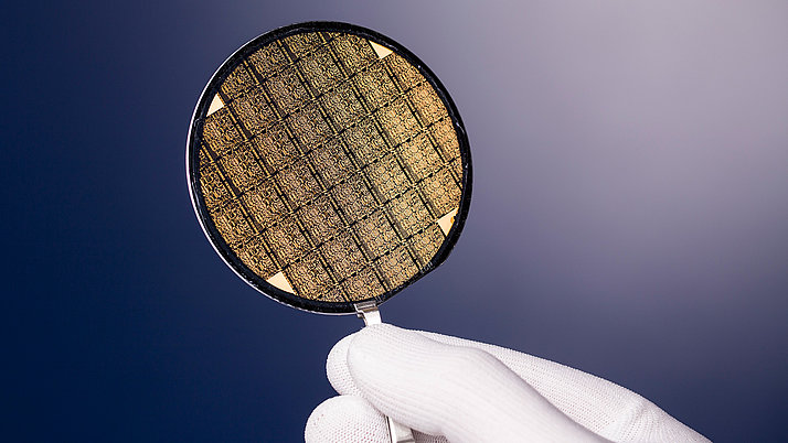 A gloved hand holds a shiny, circular silicon wafer that features a grid of intricate patterns. 