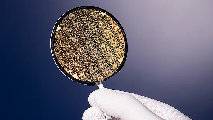 Person hält einen silbernen Halbleiter-Wafer in der Hand. Der Wafer hat eine komplexe, goldene Struktur mit vielen kleinen quadratischen Mustern.