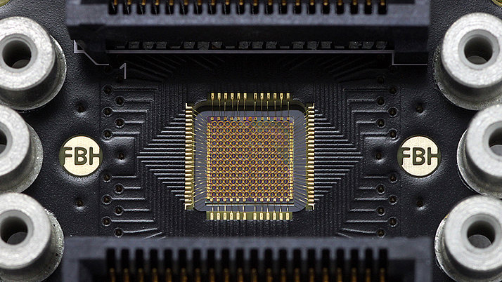 Circuit board featuring a central microchip surrounded by multiple connection ports. The microchip has a grid of small pins and is flanked by solder points and labeled areas. 