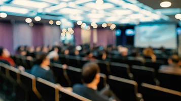 Eine große Gruppe von Menschen sitzt in einem Konferenzsaal. Sie sind von hinten zu sehen und scheinen einer Präsentation zuzuhören, die auf einer Leinwand gezeigt wird.
