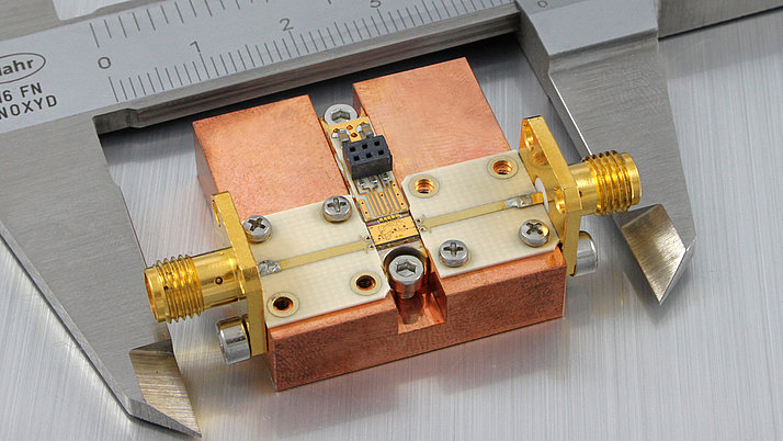 A precision electronic component is positioned under a caliper ruler. The component features a copper base with distinctive gold and silver connectors, and a small circuit board in the center.