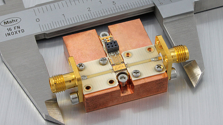 A precision electronic component is positioned under a caliper ruler. The component features a copper base with distinctive gold and silver connectors, and a small circuit board in the center.