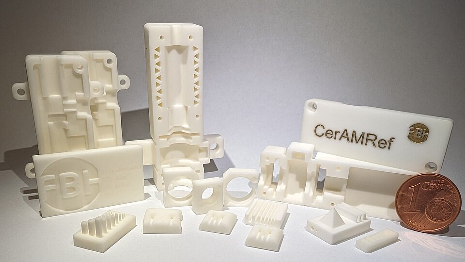 The image shows a collection of white 3D-printed ceramic parts in various shapes and sizes. Some are flat with engraved logos, while others have more complex structures. A cent coin serves as a scale for the size of the parts.