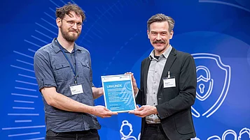 Zwei Männer stehen auf einer Bühne und halten gemeinsam eine Urkunde. Der linke Mann trägt ein graues Hemd und hat eine Brille, der rechte Mann hat einen Schnurrbart und trägt einen blauen Anzug.