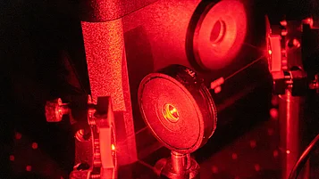 Close-up of a quantum physics experiment setup in the laboratory: a red laser beam hits several optical components such as lenses, mirrors, and mounts, which are mounted on an optical experiment plate. The metal components and adjustment screws reflect the intense red light as the laser beam passes through the optical elements.