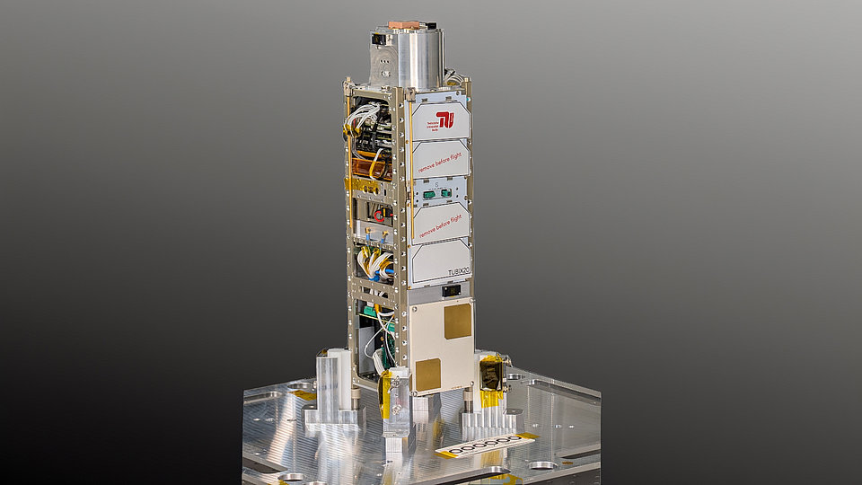 Das Bild zeigt ein hochkomplexes Gerät mit einer schlanken, stabilen Struktur, das mit verschiedenen elektronischen Komponenten und Drähten ausgestattet ist. Es steht auf einer metallischen Basis, die eine solide Unterstützung bietet. Der Hintergrund ist grau und hebt das Gerät hervor.