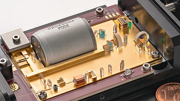 Nahaufnahme eines offenen Quantenlicht-Lasermoduls, das im Inneren eine goldfarbene Platine zeigt mit verschiedenen optischen Komponenten, einem großen silbernen Element, transparenten Linsen und weiteren mikroelektronischen Bauteilen. Ein Centstück ist als Größenvergleich im Bild platziert.