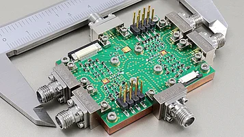 Photograph shows the demonstrator module with the full-duplex topology, comprising two power amplifiers and a low-noise amplifier combined with a printed circuit board, which is mounted onto a copper heat sink. The module is placed between the jaws of a caliper for size reference.